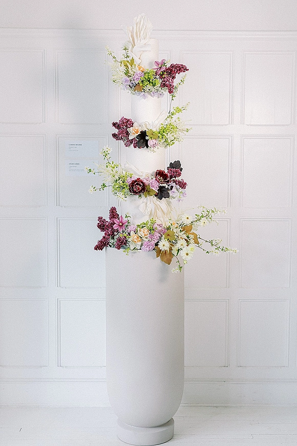 Wedding cake with sugar and fresh flowers on a pedestal, four-tier white buttercream design with greenery against a paneled wall