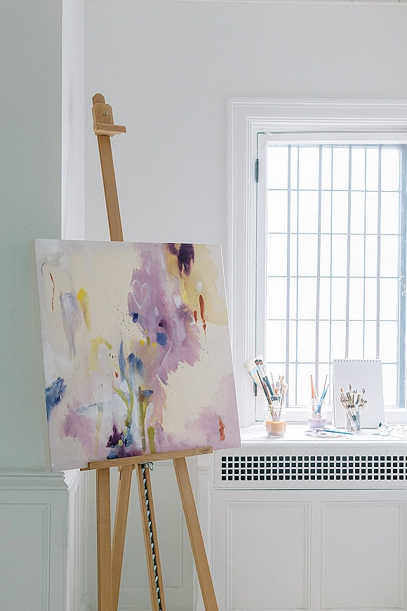 Wedding guest book on an easel with a canvas painting, paintbrushes in jars, and pens by a large window on a white wall backdrop