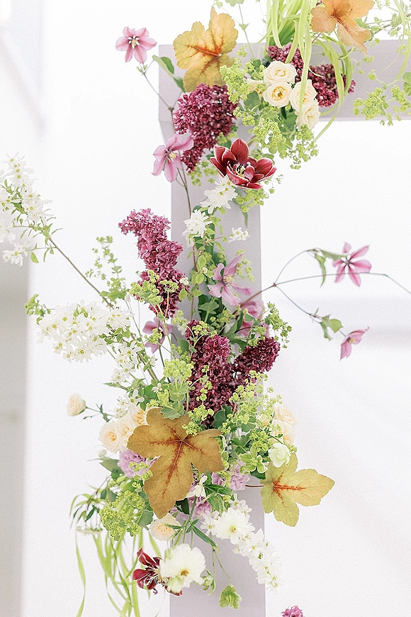 Wedding floral arrangement with lilac blooms, roses, and trailing greenery on a wooden structure against a clean white backdrop