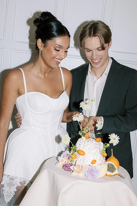 Wedding cake cutting by bride and groom, slicing a white textured cake with fresh flowers and fruit decor on a linen table indoors
