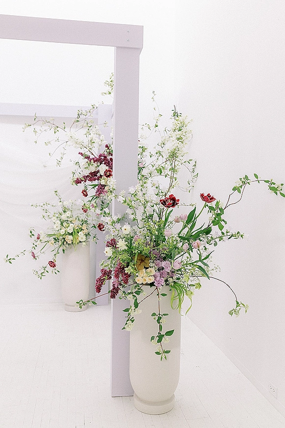 Wedding ceremony floral arrangement with wedding arch flowers, greenery vines, and white floor vases against a clean white wall backdrop