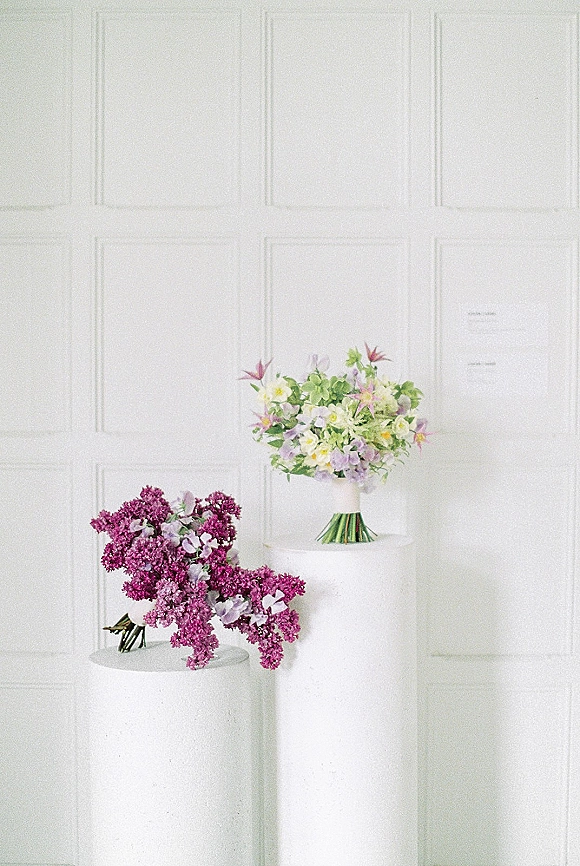 Wedding bouquet with pastel bridal bouquet tones and lilac blooms, hand-tied with ribbon wrap on white pedestal stands against paneled wall