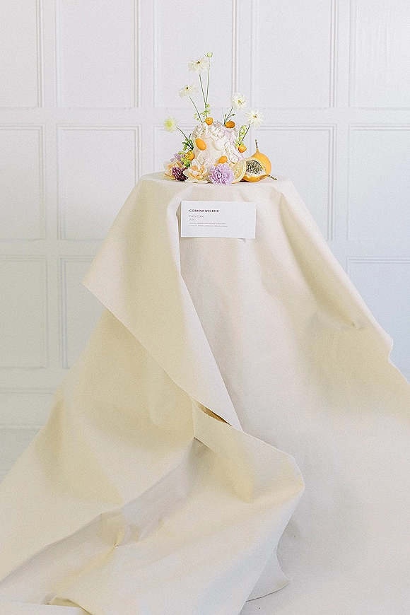 Wedding cake with fresh flowers and citrus on a linen-draped table, featuring a floral topper and place card against a white paneled wall