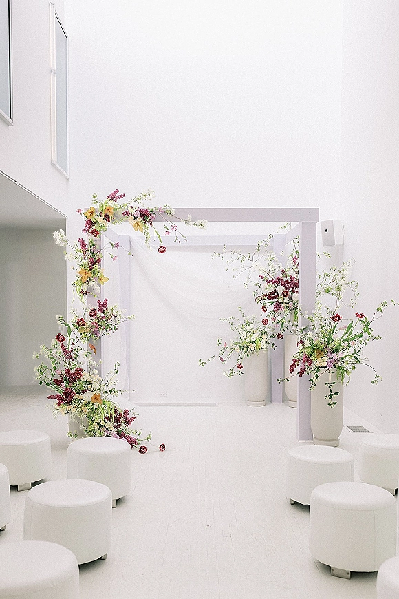 Ceremony backdrop with modern wedding altar featuring a white arch, draped fabric, and floral pedestals in a minimalist room