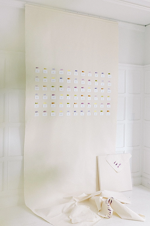 Seating chart display with wedding escort card wall of pastel paint swatch cards on paper backdrop stand in a bright white studio corner