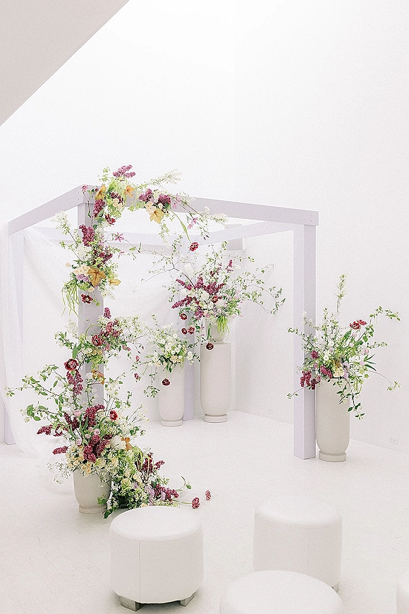 Wedding ceremony backdrop with modern ceremony altar featuring an asymmetrical floral arch, greenery, and white pedestals in a white studio space