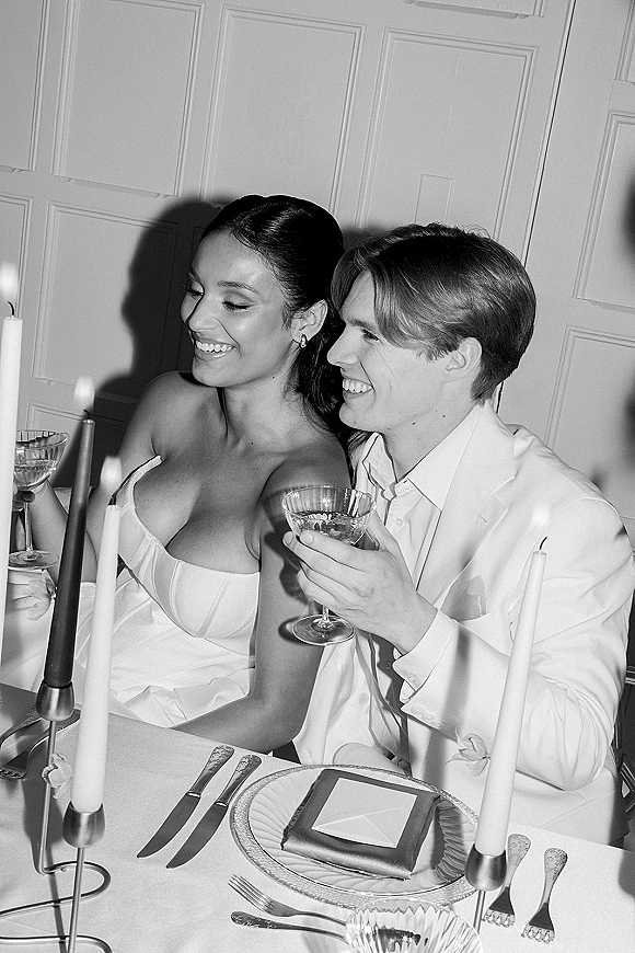 Reception couple portrait of newlyweds toasting with stemmed cocktails at a sweetheart table lit by taper candles in an indoor space