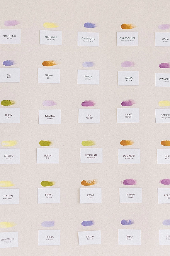 Escort card display with wedding escort cards in calligraphy and watercolor paint swatches arranged on a neutral wall
