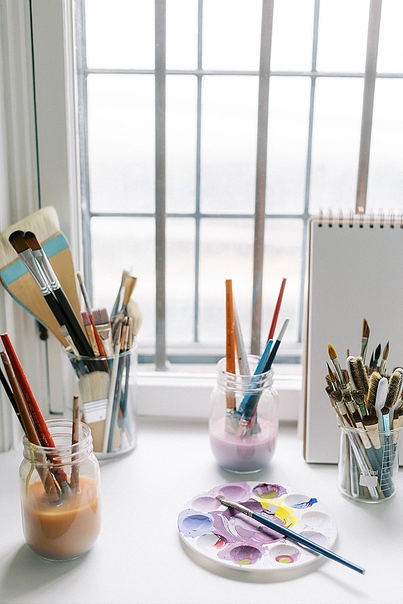 Artist workspace with painting supplies flatlay, brushes in glass jars, watercolor palette and sketchbook on a desk by a bright window