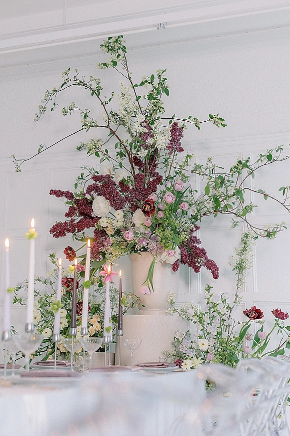 Wedding reception centerpiece with a large urn floral arrangement, flowering branches, greenery, taper candles, and place settings indoors