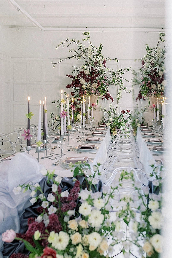 Reception tablescape with long banquet table decor featuring floral centerpieces, tall taper candles, gray napkins and clear acrylic chairs in a white room