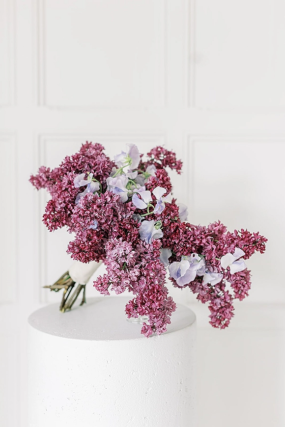 Bridal bouquet of lilac wedding bouquet and sweet peas with ribbon wrap on a white pedestal against a white paneled wall backdrop