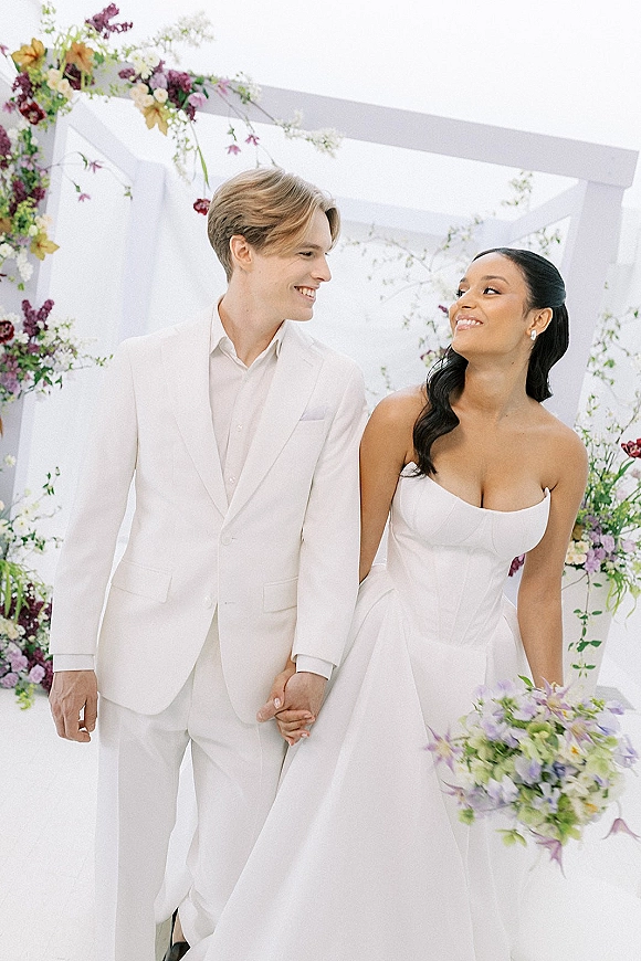 Couple portrait of bride and groom holding hands, smiling under a floral arch with bridal bouquet and white draped backdrop