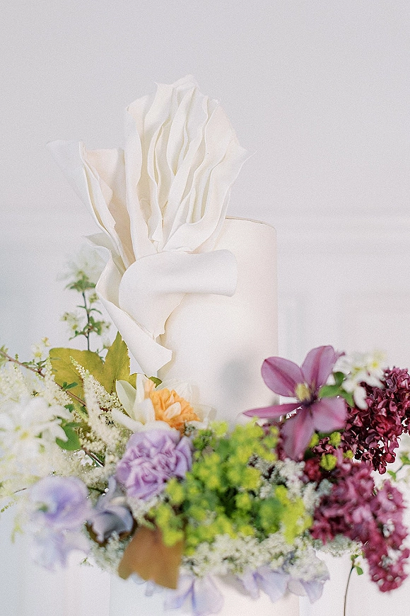 Wedding cake with white buttercream cake finish, sculptural wafer paper ruffles, and fresh purple and green florals against a white backdrop