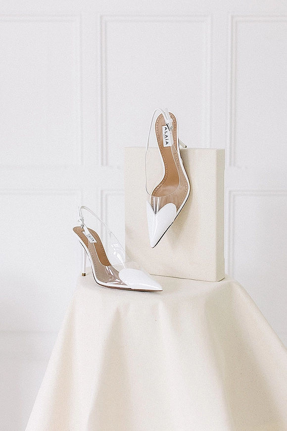 Bridal shoes, sleek white slingback wedding heels with clear PVC straps and pointed toes on a shoebox against a white paneled wall