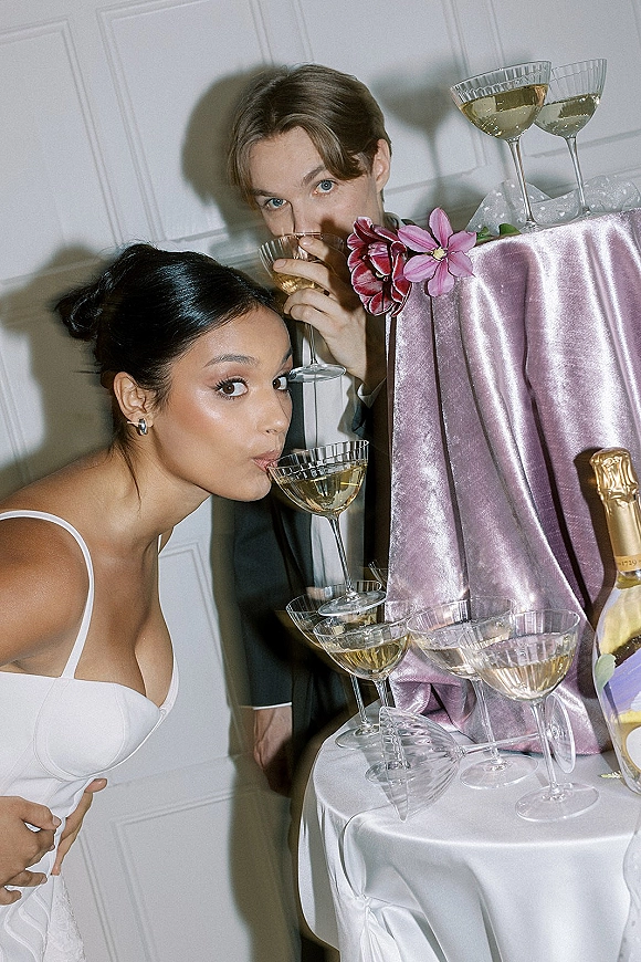Wedding toast moment with bride and groom toast sipping champagne beside coupe glasses on satin table with pink drape against white paneled wall