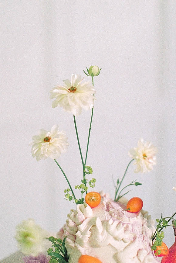 Wedding cake with buttercream frosting, piped texture, edible flowers and kumquats with greenery against a light gray backdrop