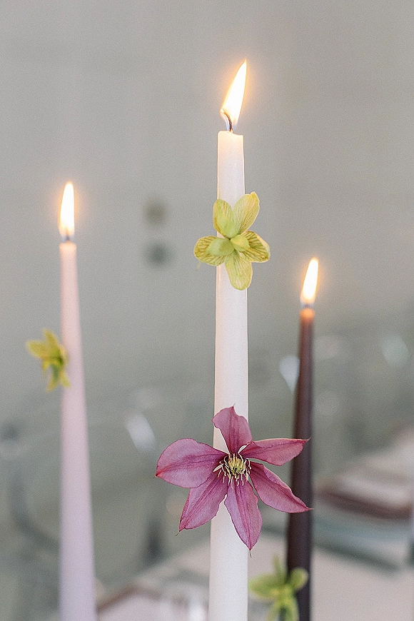 Wedding taper candles in white, blush, and brown with floral adornments and flickering flames on a blurred reception table with glassware