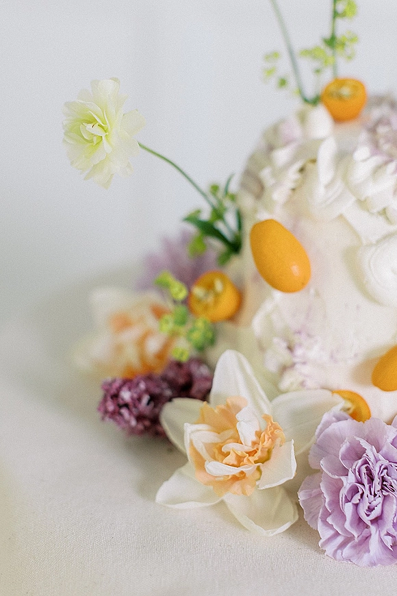 Wedding cake with floral wedding cake styling, white buttercream frosting topped with sugar flowers, fresh blooms, and kumquats on a white tablecloth