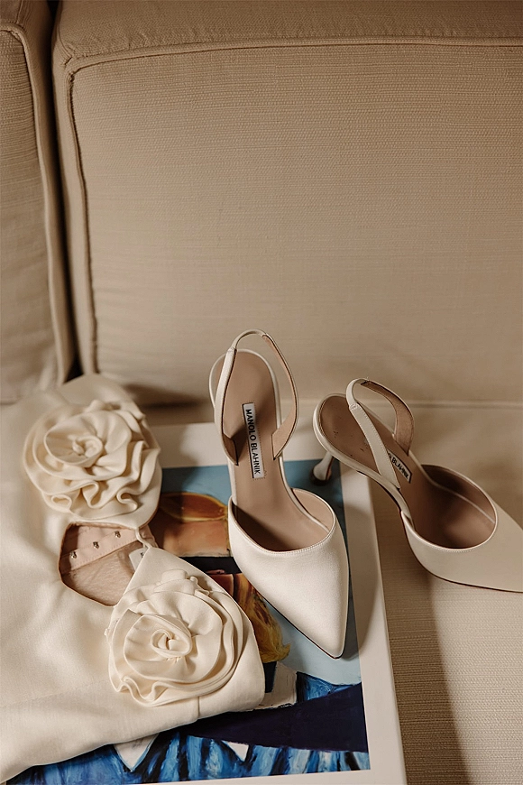 Wedding shoes flat lay of white slingback heels with rosette embellishments on a shoe box and printed art book atop a beige chair