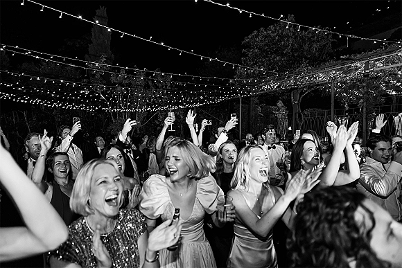 Wedding reception dance floor packed with guests cheering in formalwear under string lights on a garden patio at night