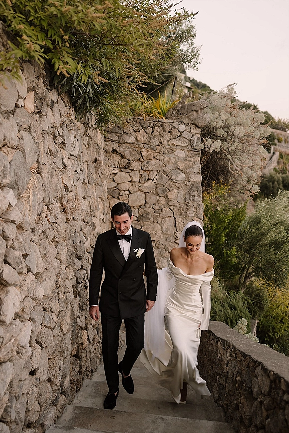 Wedding couple portrait of bride and groom walking hand in hand up outdoor stone stairs, veil flowing behind her off-shoulder gown train