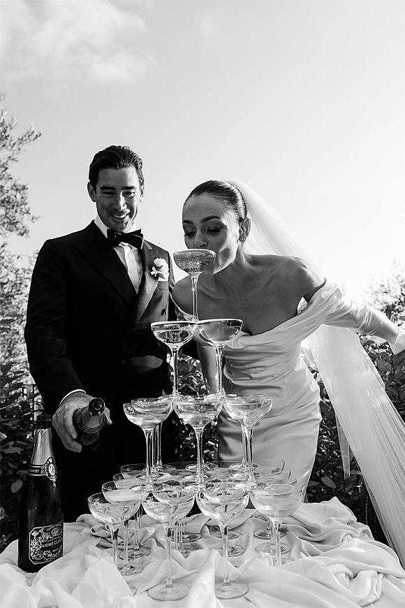 Champagne tower moment as bride in veil and gloves sips from coupe glasses while groom in tux pours, outdoors under trees and sky