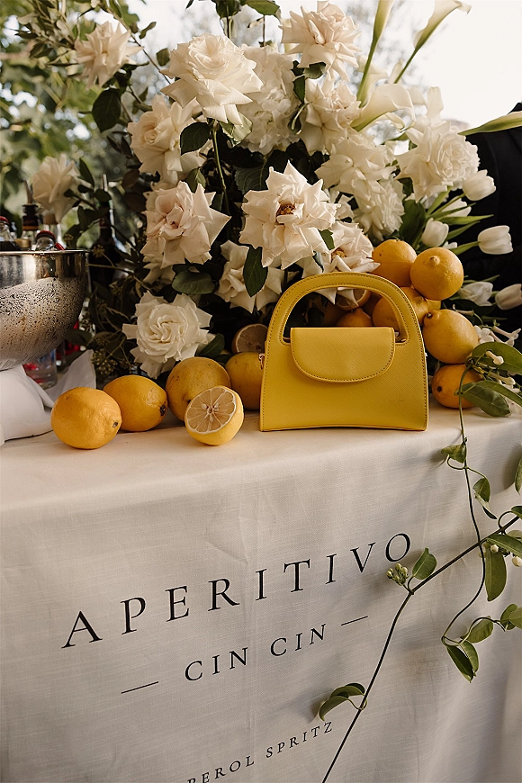 Wedding bar cart with Aperol spritz wedding bar sign, lemons and citrus slices, and white roses and tulips on linen outdoors in daylight