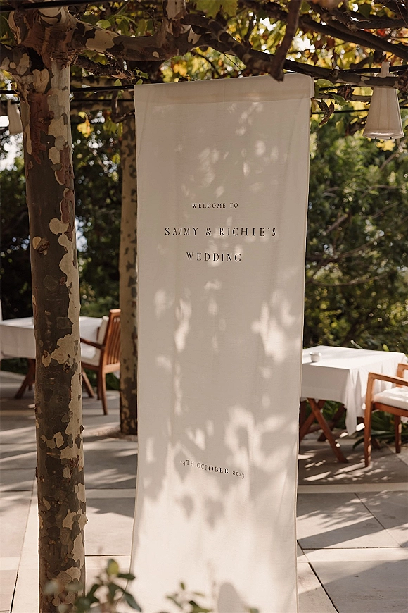 Wedding welcome sign fabric banner with black lettering hanging from a tree under leafy canopy on an outdoor patio with lanterns and tables