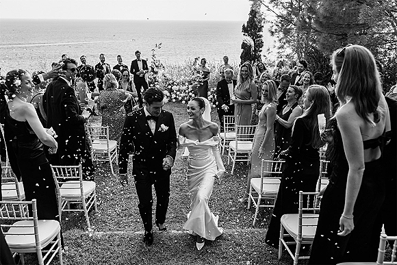 Wedding recessional with confetti wedding exit as bride and groom walk the aisle, guests cheering under a floral arch with ocean view backdrop