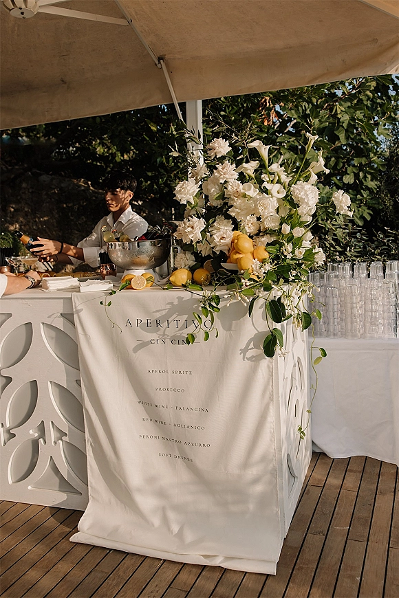 Wedding bar setup with an outdoor wedding bar, featuring a white floral arrangement and lemons on the counter under a canopy tent on a wooden deck