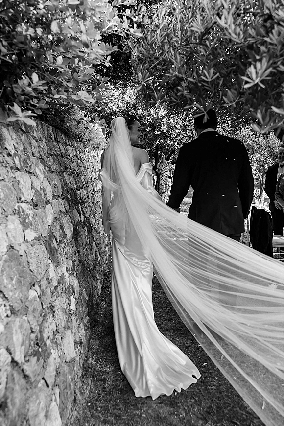 Wedding processional with bride walking down aisle hand in hand with groom, long veil flowing along a garden path under tree canopy