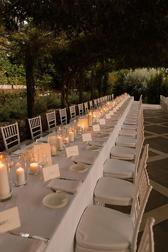 Reception tablescape with a long banquet table wedding setup, white tablecloth, chiavari chairs, pillar candles, and place cards on an outdoor terrace