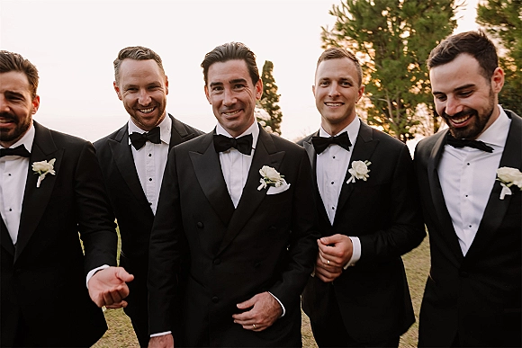 Groomsmen portrait of groom with groomsmen in black tuxedos and bow ties, standing on a lawn with trees in warm sunset light