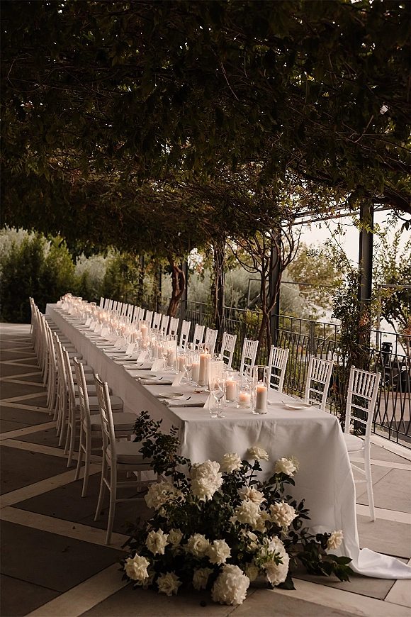 Reception tablescape with long banquet table dressed in white linens, taper and pillar candles, and white florals on an outdoor terrace under trees