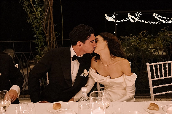 Wedding kiss portrait of bride and groom kiss at an outdoor patio reception table under string lights, with wine glasses on white tablecloth