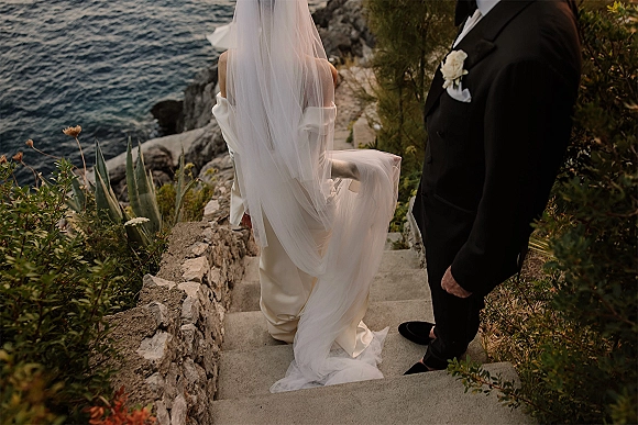 Wedding couple portrait from behind on stone steps, bride’s long veil train flowing beside groom in tuxedo by the ocean cliffs.