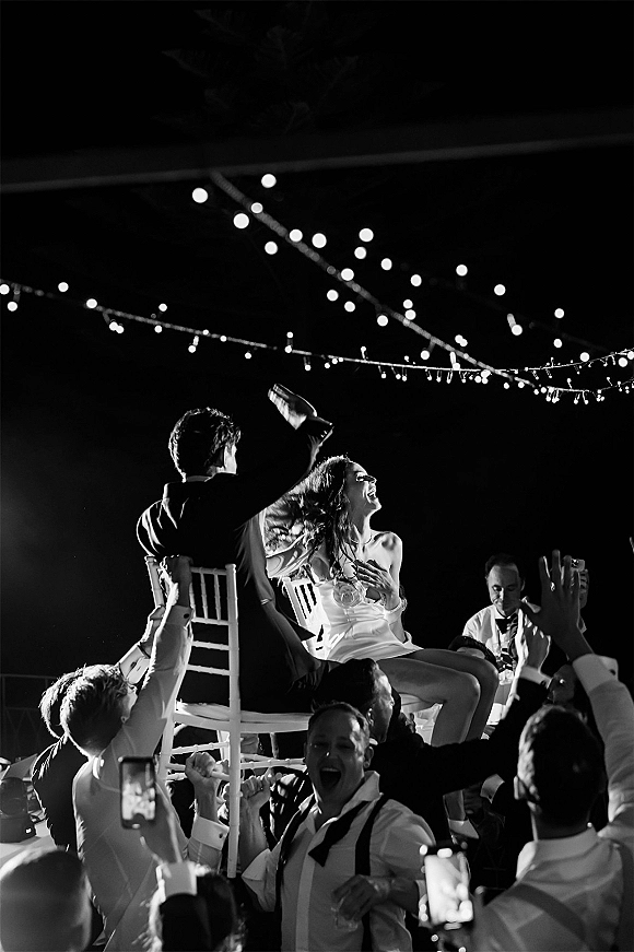 Wedding chair lift during horah chair dance as guests raise bride and groom on chairs under string lights at an outdoor night reception