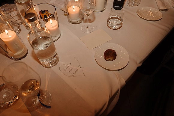 Reception tablescape with candlelit wedding table details, pillar candles in glass hurricanes, wine glasses, plates, and handwritten note card in dim light
