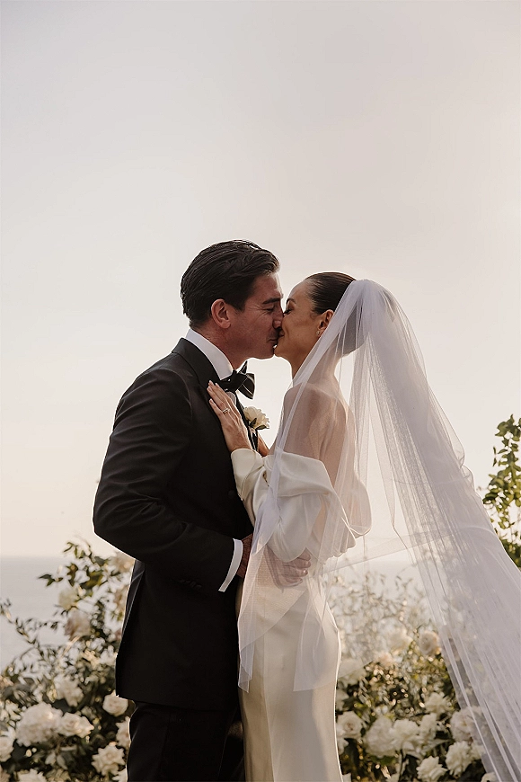 Wedding kiss portrait of bride and groom kiss, her long veil blowing at sunset with ocean horizon behind, groom in black tuxedo and bow tie