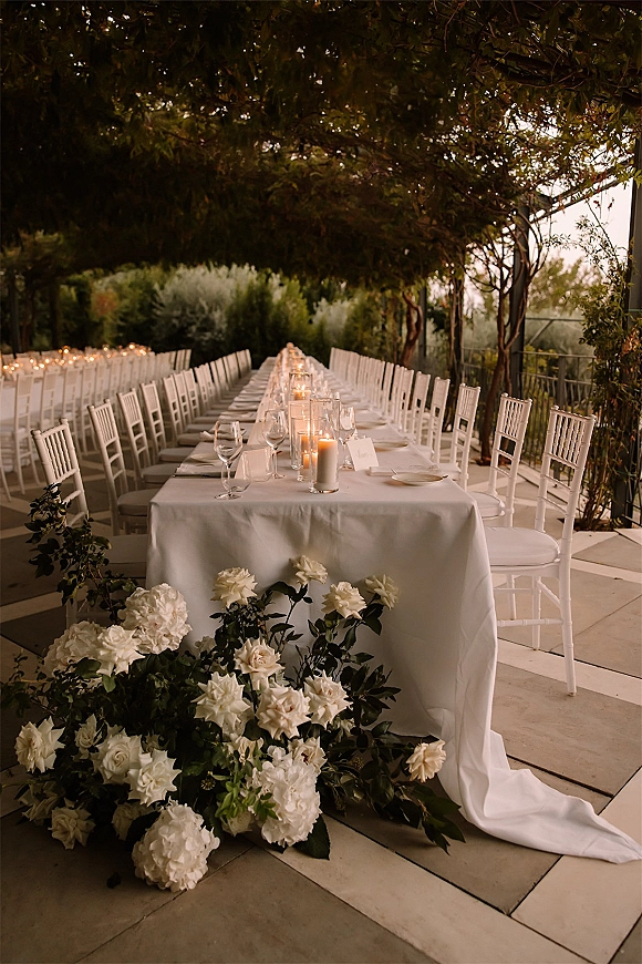 Reception tablescape with a long banquet table wedding setup, pillar candles in glass hurricanes, white florals and greenery on a garden patio