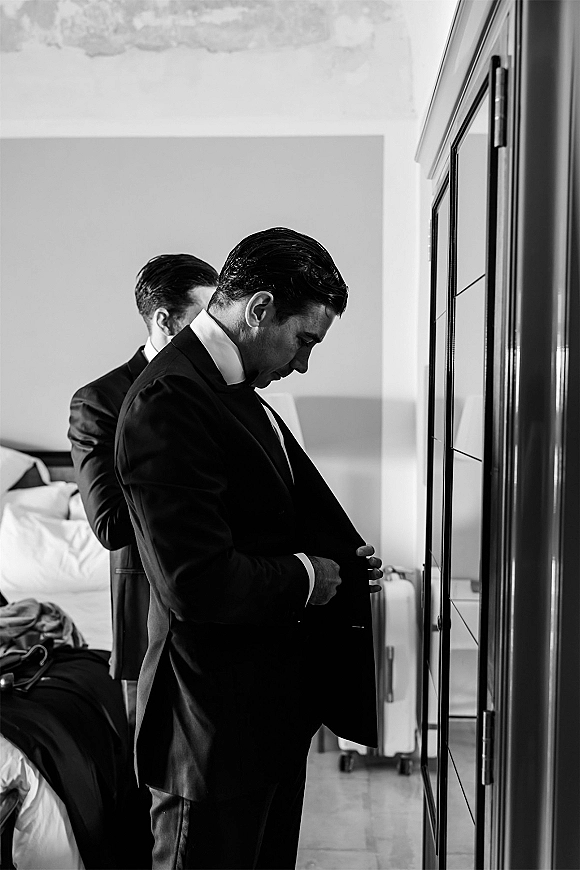 Groom getting ready as he puts on his suit jacket over a dress shirt in a bedroom with wardrobe and luggage behind him