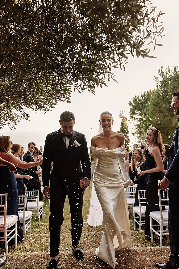Wedding recessional as newlyweds walk down the outdoor aisle holding hands while guests toss confetti, bride in veil and groom in tuxedo