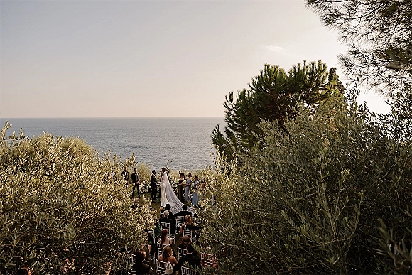 Wedding ceremony with bride in long veil and groom at a white floral arch, guests seated on chiavari chairs, ocean view behind