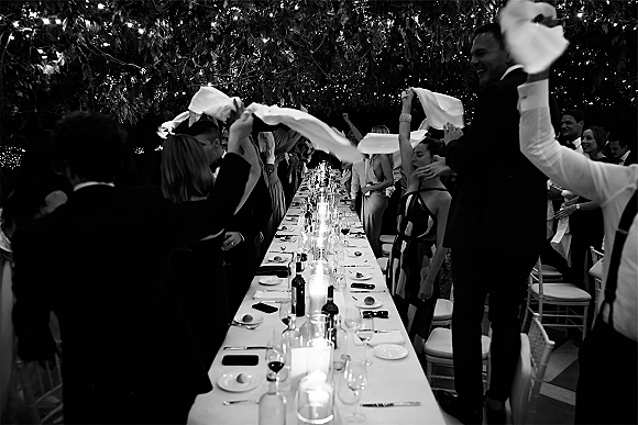 Wedding reception toast as guests cheer around a long banquet table with candlelight and white linens under string lights in trees at night