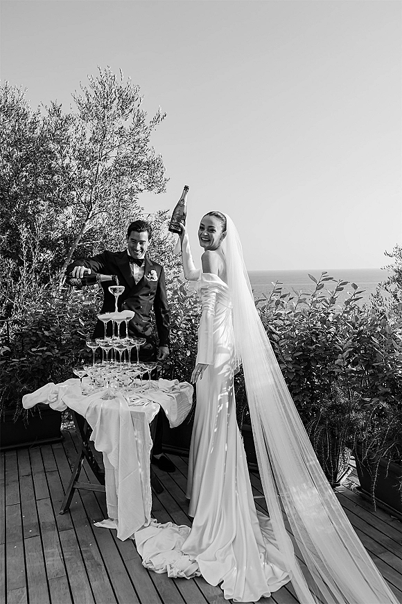 Champagne tower moment as bride and groom pour bottles into stacked coupe glasses, her long veil flowing on an ocean-view deck