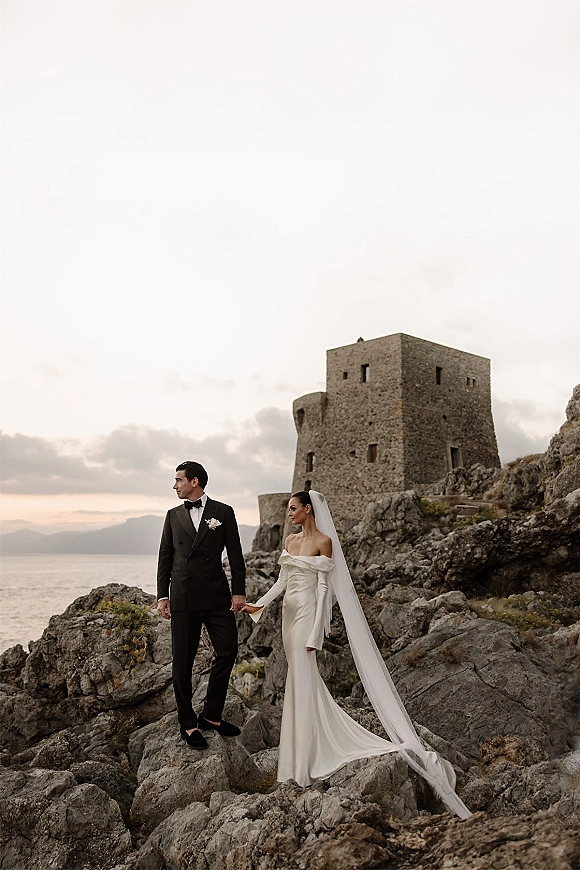 Couple portrait of bride and groom holding hands, her long veil in the wind by a stone castle on a rocky coastline above the ocean