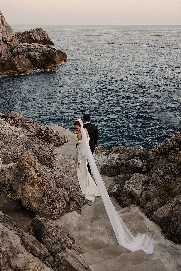 Couple portrait on a cliffside wedding portrait with the bride’s long veil trailing down stone stairs above the rocky ocean horizon
