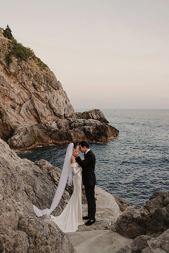 Wedding couple portrait in a cliffside wedding portrait, bride in long sleeve dress with veil and groom in tuxedo embracing on rocky coast by ocean cliffs