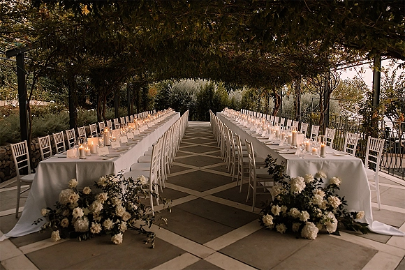 Reception tablescape for an outdoor wedding reception with long banquet tables, white florals and greenery garlands, and glowing taper candles beneath trees